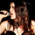 Female singer passionately singing into a microphone on stage, dark background behind her.