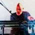 Bald male musician on a keyboard synth, singing into a microphone amid stage smoke.
