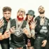 Six adults in edgy metal/punk outfits posing for a group photo, some showing hand signs and tattoos, against a light background